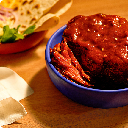 Bowl of barbecued meat on a wooden table with a sandwich in the background