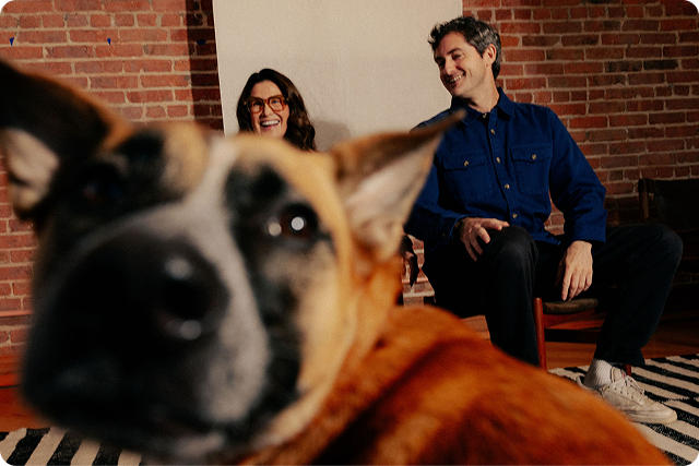 Two people sitting on a couch with a dog in the foreground, in a room with brick walls.