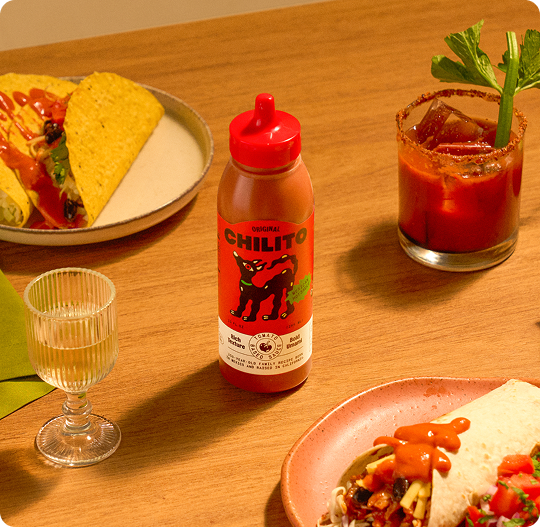 Table with plates of food, a bottle of Chilito hot sauce, and a Bloody Mary.